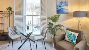 Ein modernes Wohnzimmer mit Glastisch, zwei Stühlen, einer Couch mit Zierkissen, einer Stehlampe und einem großen Fenster mit Jalousien.