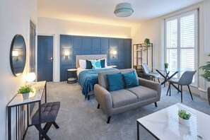 A modern bedroom with a large bed, a sofa, a dining table, and a chair. There is a mirror, a lamp, and a plant on the side table. - Host & Stay | The Langley Hotel, Apartment 5 (Whitby)