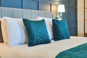 Una cama con almohadas blancas y turquesa, una manta turquesa y una lámpara fijada a la pared.