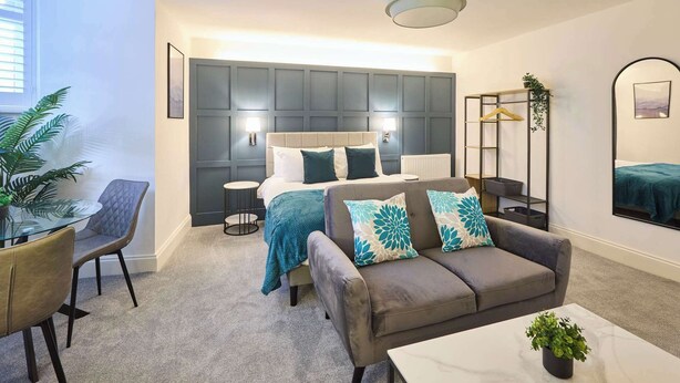 Ein modernes Schlafzimmer mit einem großen Bett, einem Sofa, einem kleinen Tisch und einem Sessel. Auf dem Tisch steht eine Pflanze und an der Wand hängt eine Hängepflanze.