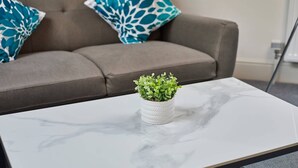 Una mesa de centro de mármol blanco con una pequeña planta en maceta en el centro, ubicada frente a un sofá gris con dos cojines estampados.