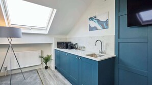 Cocina pequeña con armarios azules, encimera blanca, un lavamanos y un microondas. Hay un tragaluz sobre la ventana, una planta en maceta y un televisor de pantalla plana montado en la pared.