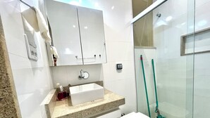 Ein modernes Badezimmer mit einem weißen Waschbecken, einem Spiegel und einer Toilette. Eine Duschkabine aus Glas und ein grüner Besen, der an der Wand lehnt.