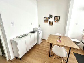 Uma cozinha compacta com armários brancos, micro-ondas e uma pequena geladeira. Há uma mesa de jantar de madeira com duas cadeiras e um vaso de planta. As paredes são decoradas com três obras de arte emolduradas.