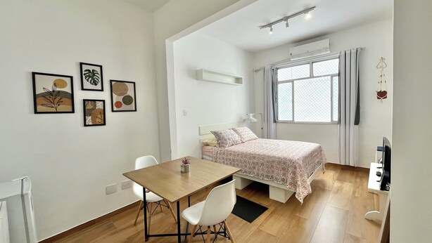 Ein kleiner, gut beleuchteter Raum mit einem Bett, einem Tisch und Stühlen. Drei Bilder in Rahmen hängen an der Wand.