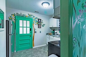 A kitchen with a green door, white cabinets, and a wall mural of a tree with fruit.