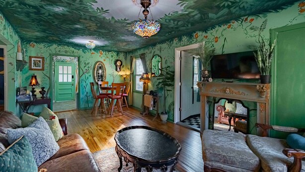 Une pièce avec un mur vert, un plancher en bois, un canapé en cuir marron, un fauteuil à motifs, une petite table ronde et un téléviseur posé sur un meuble.