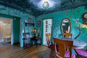 A dining area with a round table, chairs, a chandelier, and a wall with floral and tree designs.