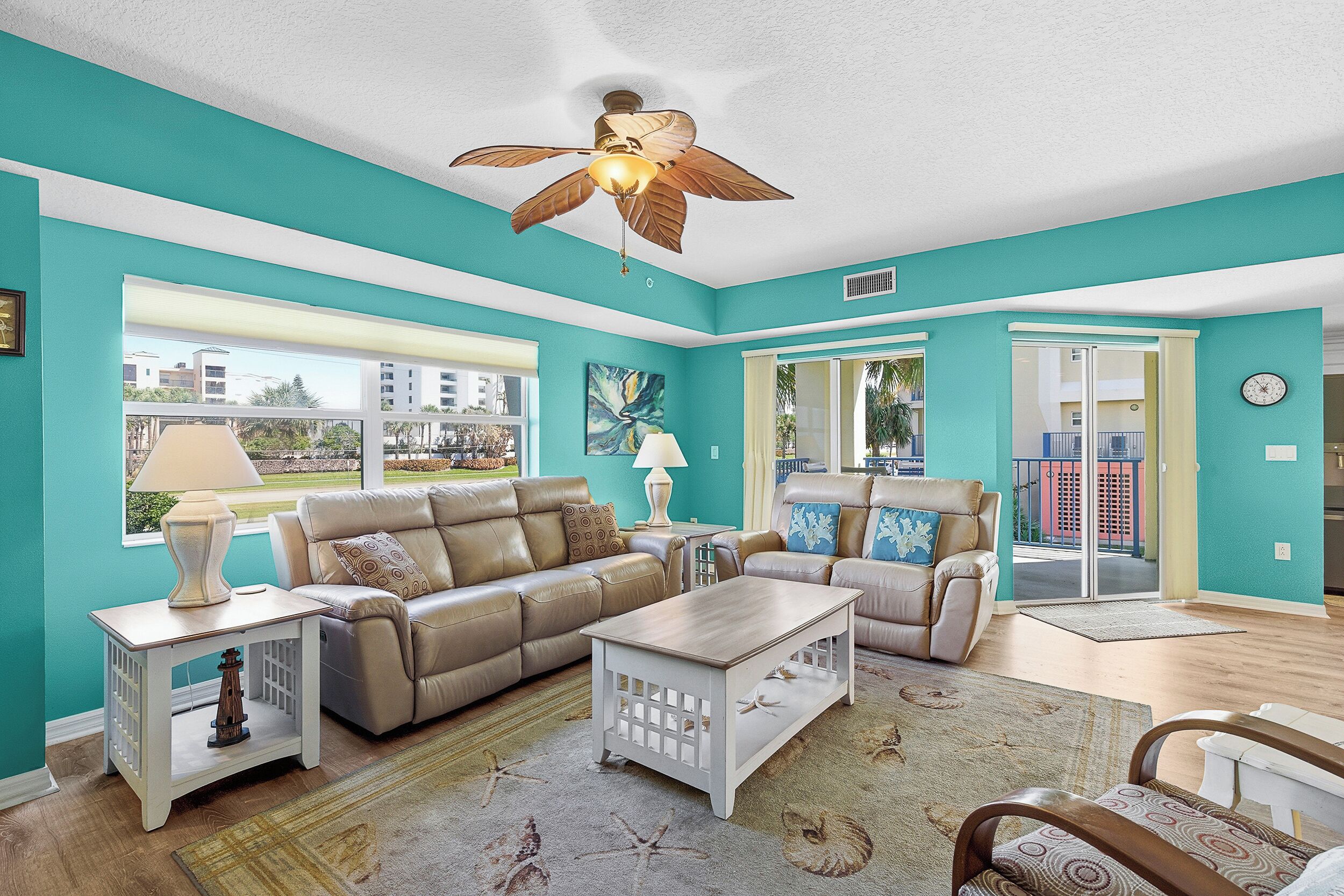 A living room with teal walls, a ceiling fan, a beige leather sofa, a white coffee table, and a view of the outside.