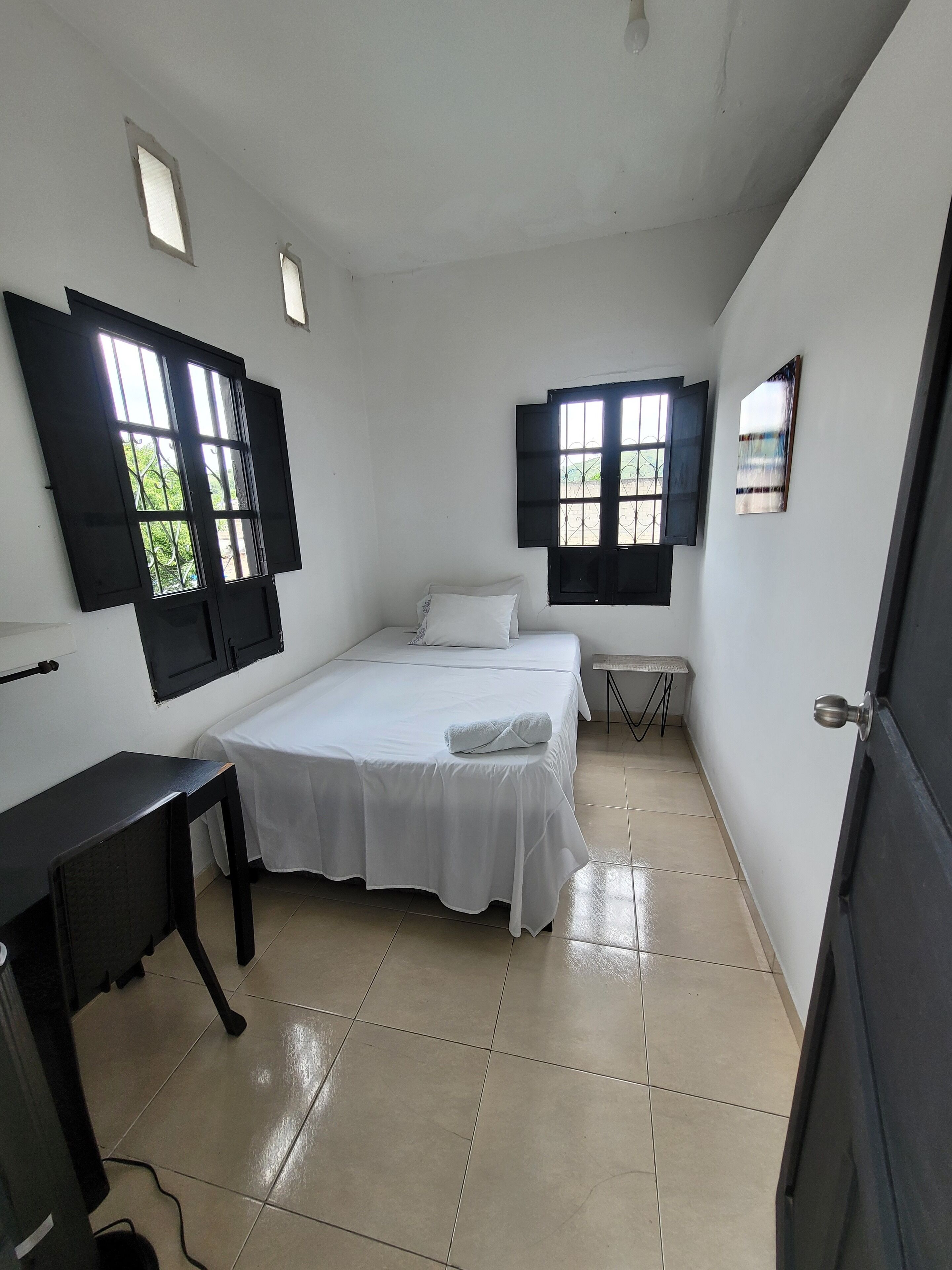 A bedroom with a bed, a desk, and two windows with shutters.