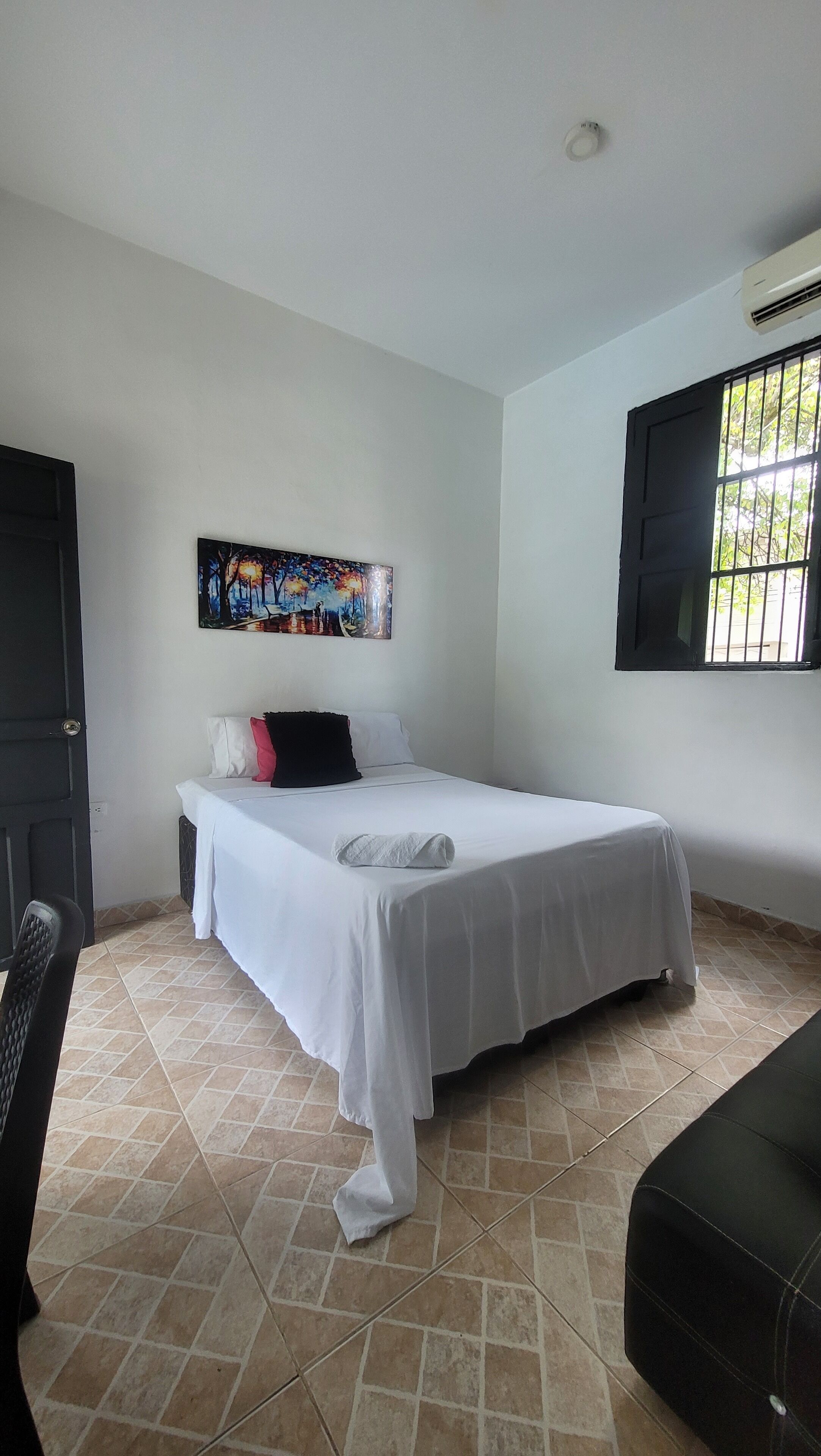A bedroom with a bed, a painting on the wall, a window with bars, and a door.