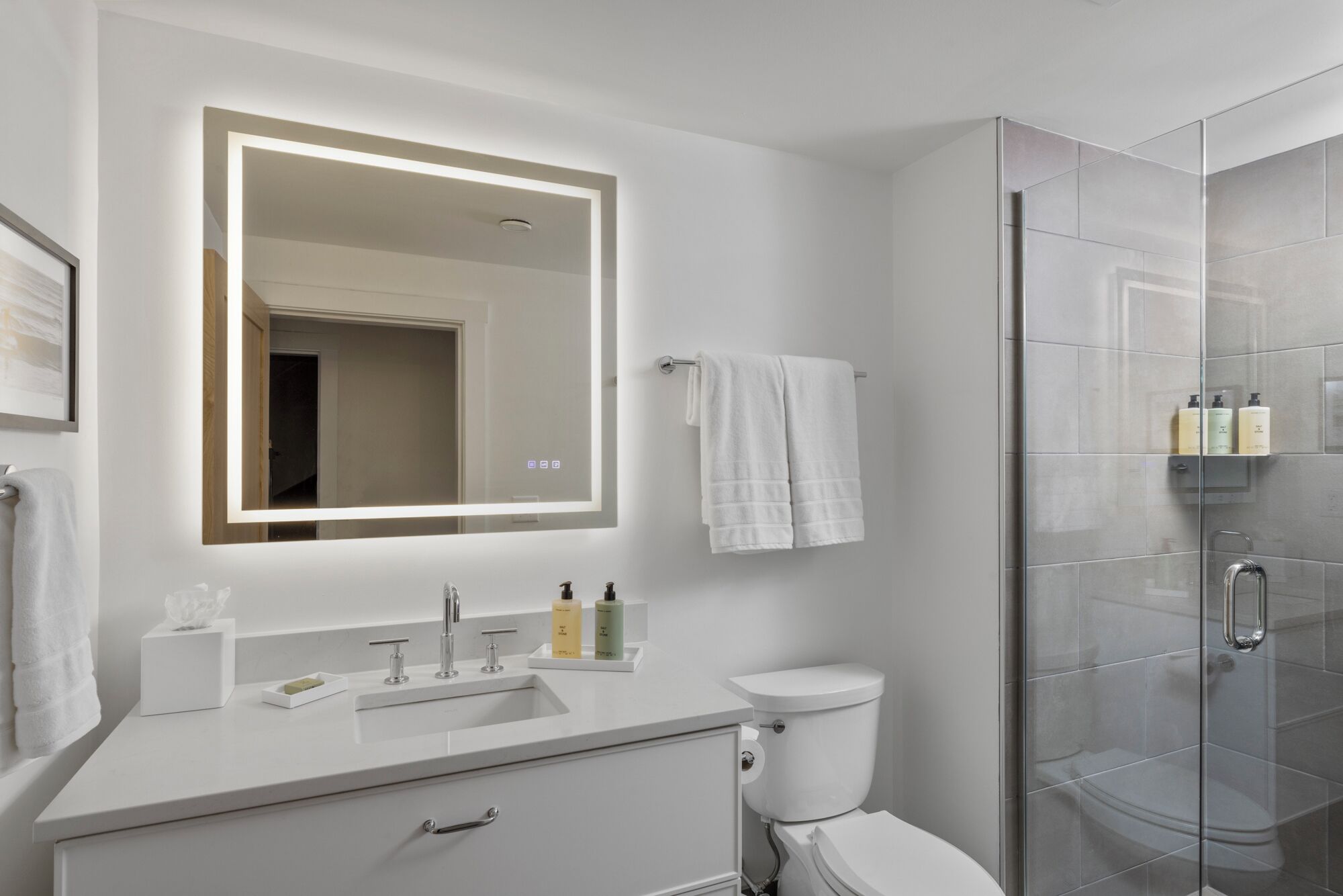 A modern bathroom with a white sink, a toilet, a glass shower enclosure, and a large mirror with integrated lighting.