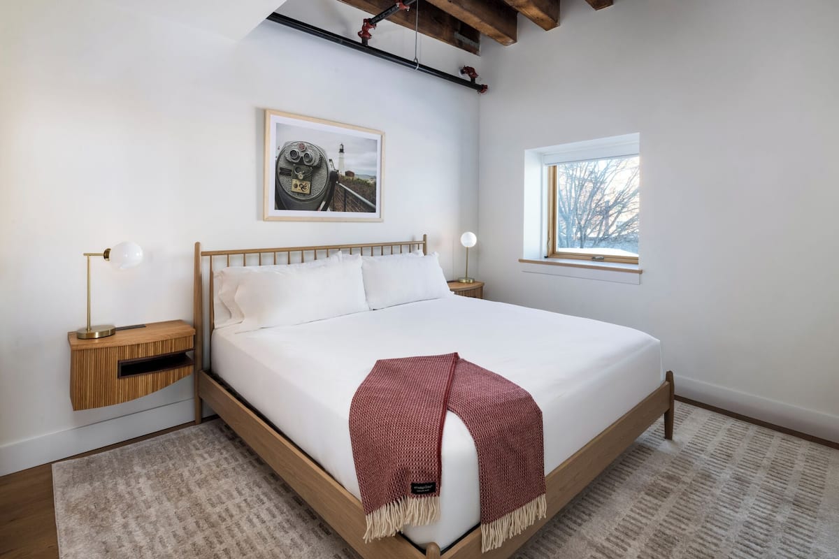 Ein Schlafzimmer mit einem hölzernen Bett, weißer Bettwäsche, einer roten Tagesdecke, einem gerahmten Bild an der Wand, einem Nachttisch mit Lampe und einem Fenster mit Blick auf Bäume.