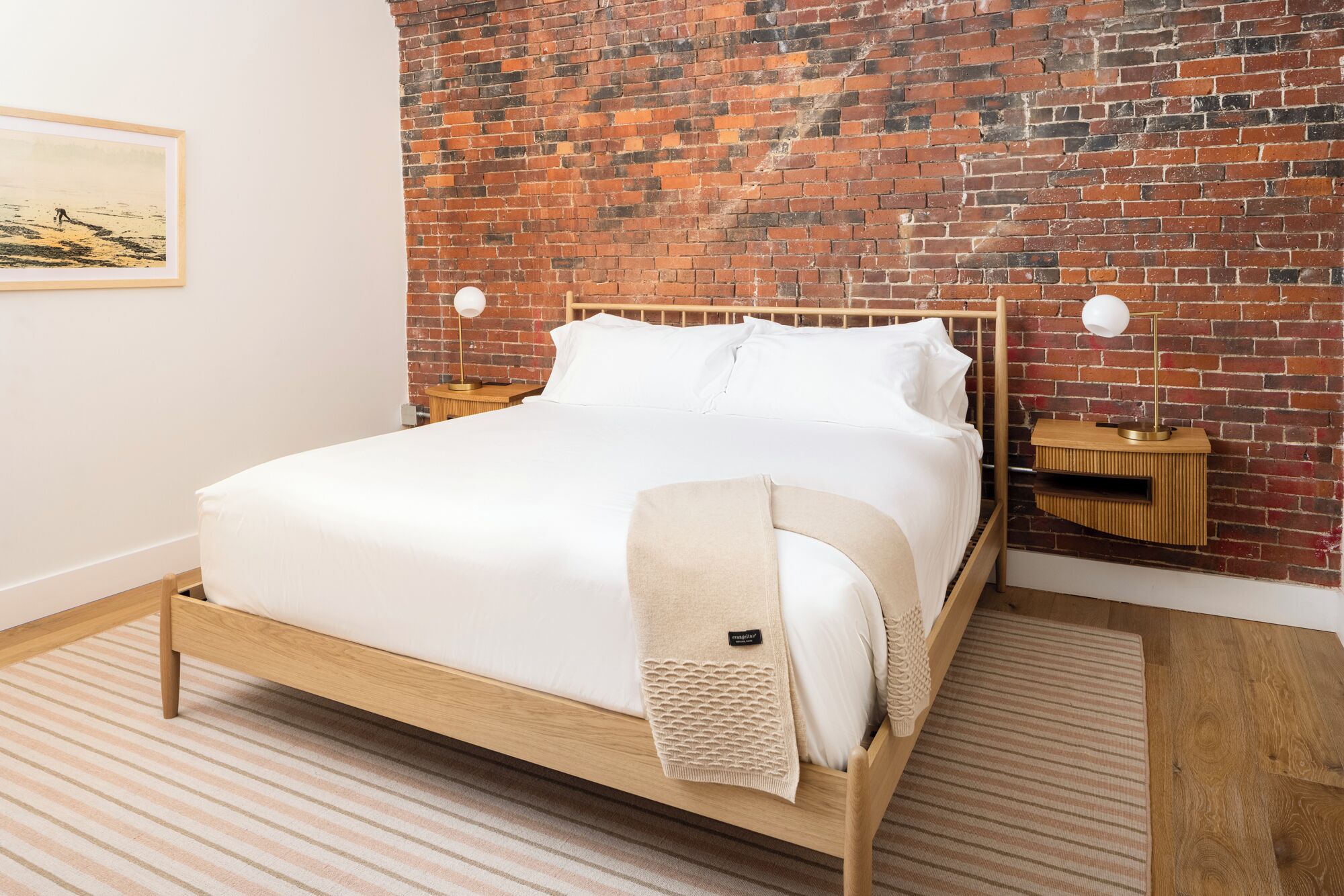 Una cama bien tendida con sábanas blancas, cabecera de madera y pared de ladrillo al fondo.