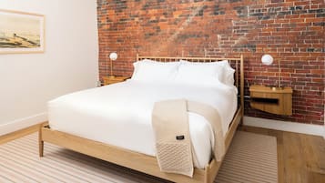 Una cama bien tendida con sábanas blancas, cabecera de madera y pared de ladrillo al fondo.