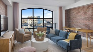 A modern living room with a large window, a TV, a sofa, a coffee table, and a dining area with a brick wall. - Evalina Hotel (Portland)