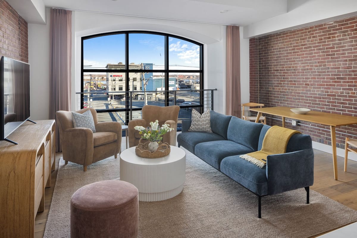 Ein modernes Wohnzimmer mit großem Fenster, einem Fernseher, einer Couch, einem Couchtisch und einem Essbereich mit Backsteinwand.