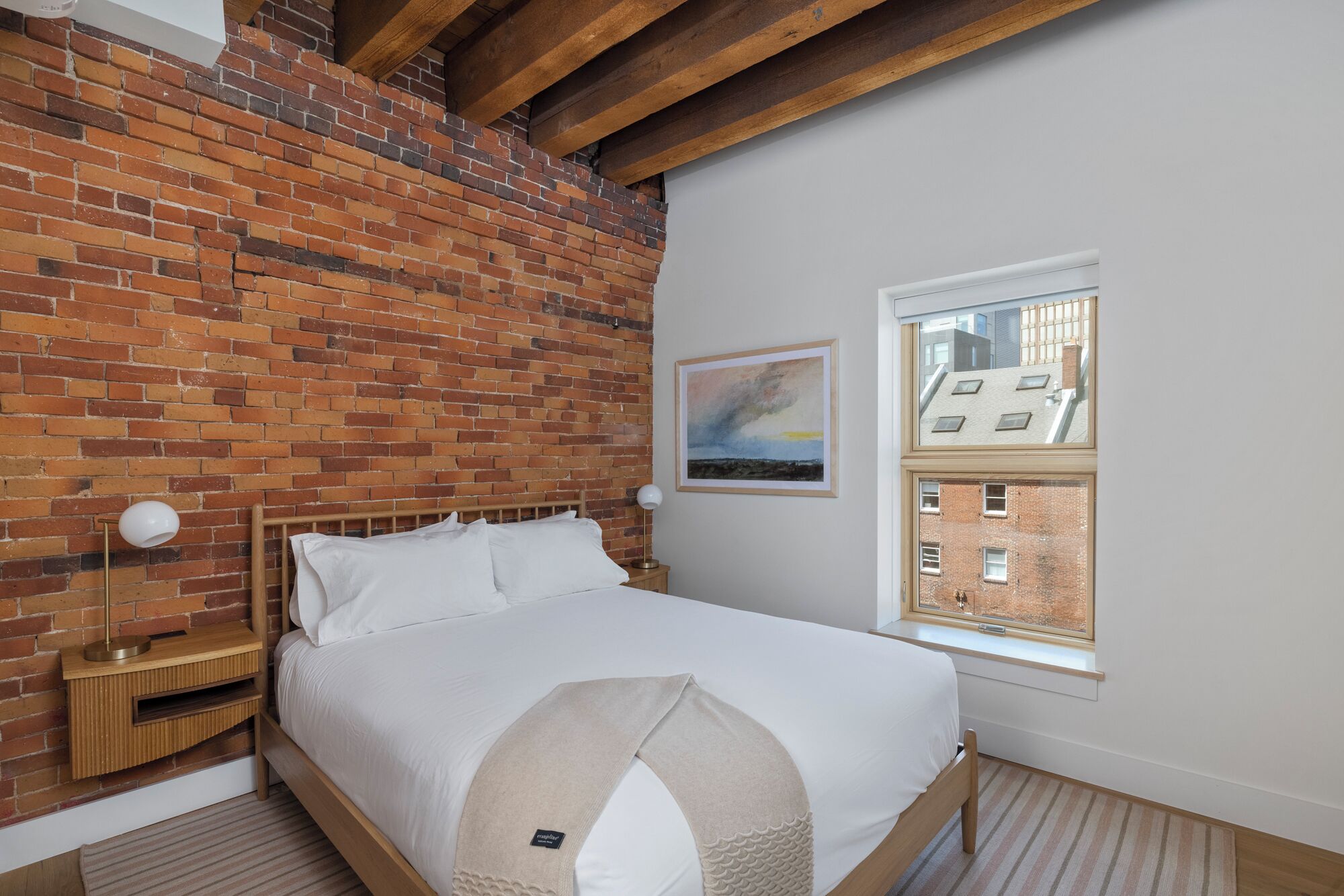 A bedroom with a brick wall, a bed with white bedding, a nightstand with a lamp, and a window with a view of buildings.