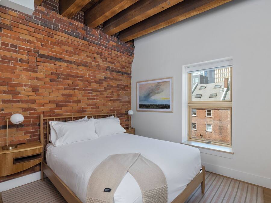 Quarto com parede de tijolos, cama com roupa de cama branca, mesa de cabeceira com abajur e janela com vista para edifícios.