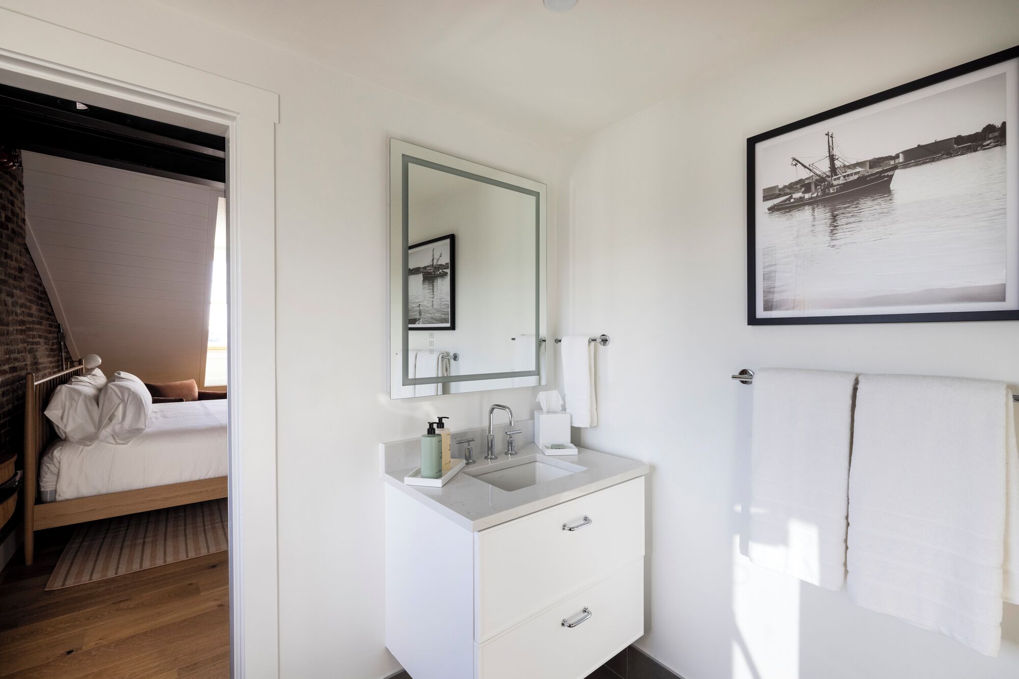 A hotel room with a bed, a bathroom with a sink, mirror, and a framed picture on the wall.