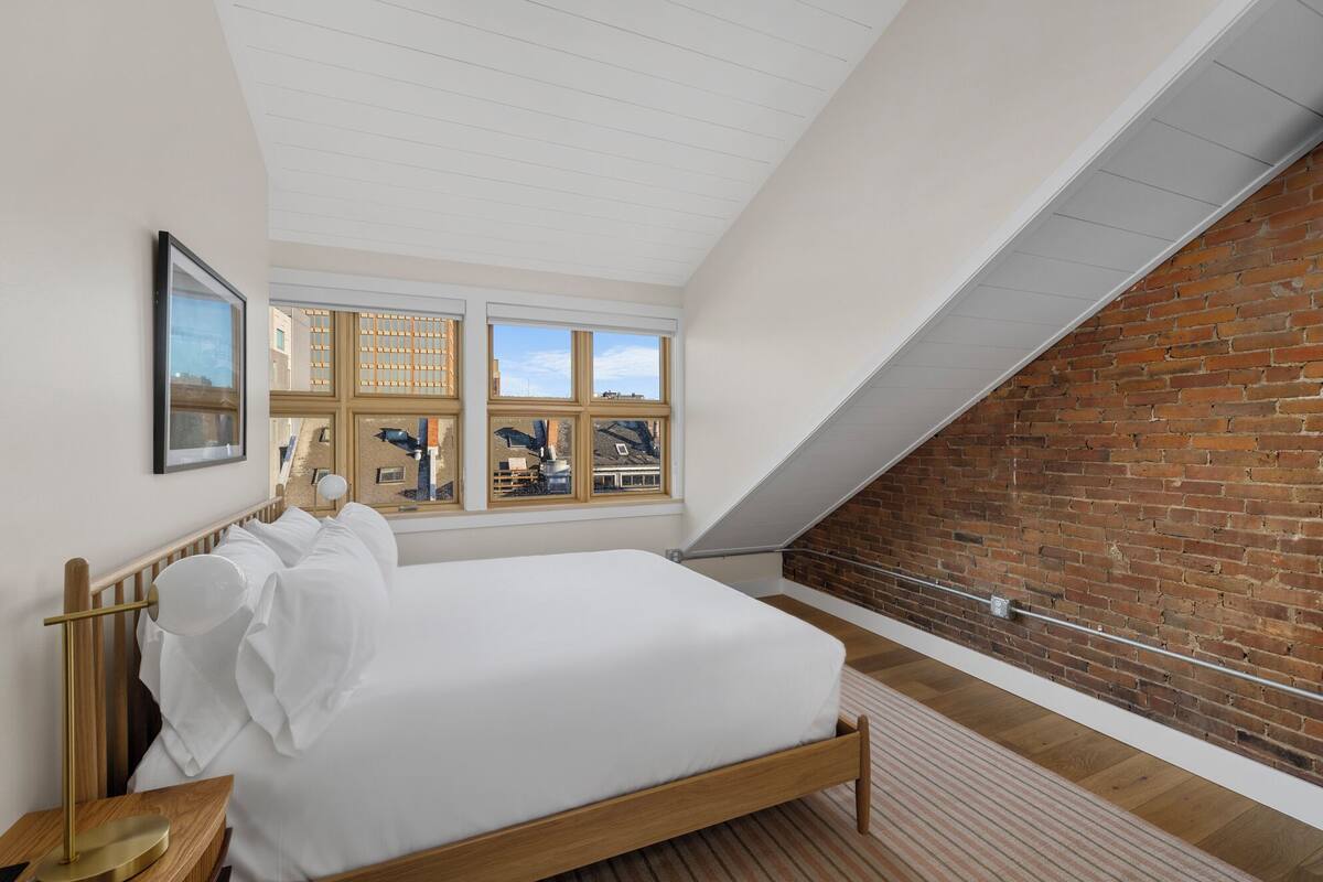 Ein Schlafzimmer mit einem hölzernen Bett, einem Nachttisch, einem gerahmten Bild und Blick auf eine Ziegelwand durch große Fenster.