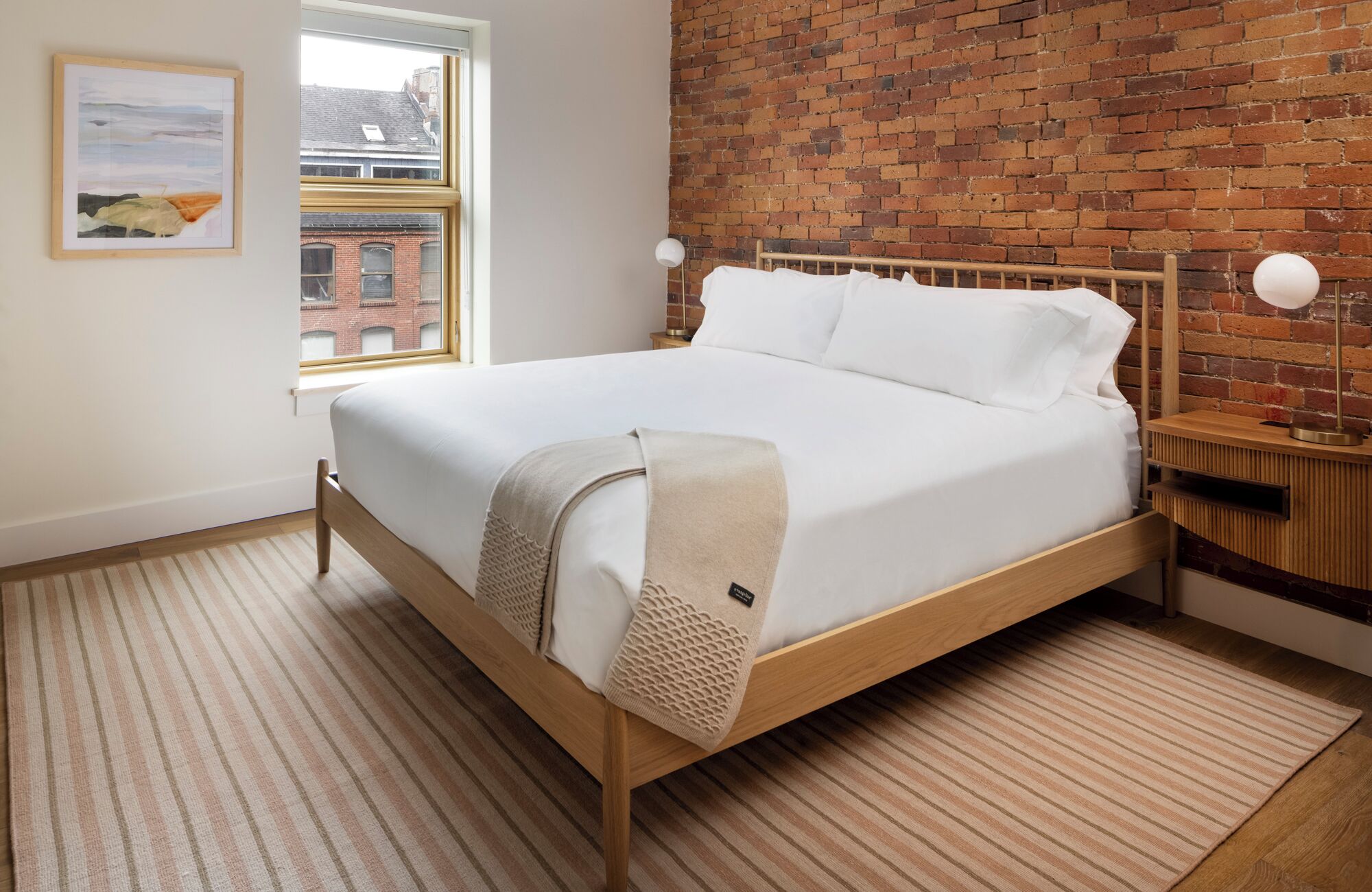 Ein Bett mit Holzrahmen, weißer Bettwäsche, einem Streifenmuster-Teppich, einer Ziegelwand und einem Fenster mit Blick auf Gebäude.