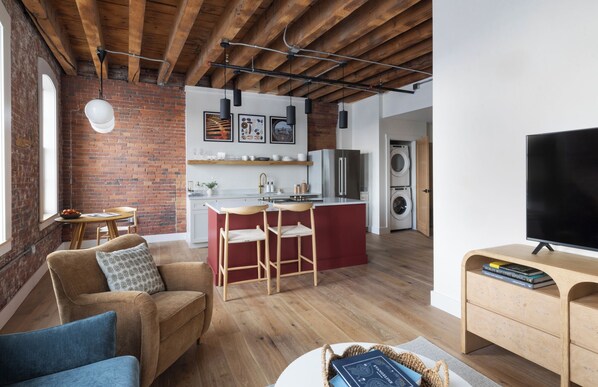 A modern living room with a brick wall, wooden ceiling beams, a sofa, a TV, and a kitchen area with a fridge and washing machine. - Evalina Hotel (Portland)