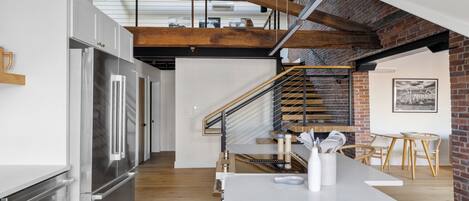 A modern kitchen with a stainless steel refrigerator, wooden beams, a brick wall, and a staircase leading to an upper level.