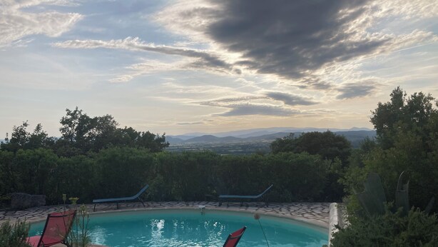 En swimmingpool med to liggestole, omgivet af stenfliser og grøn bevoksning, med udsigt til fjerne bjerge under en skyet himmel.