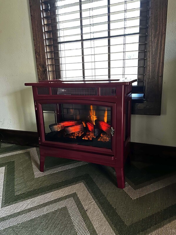 A red wood-burning stove with a glass front, positioned in a corner of a room with a patterned rug and a window with blinds. - Remote | historic cabin | renovated | Fire pit. (Rogers)