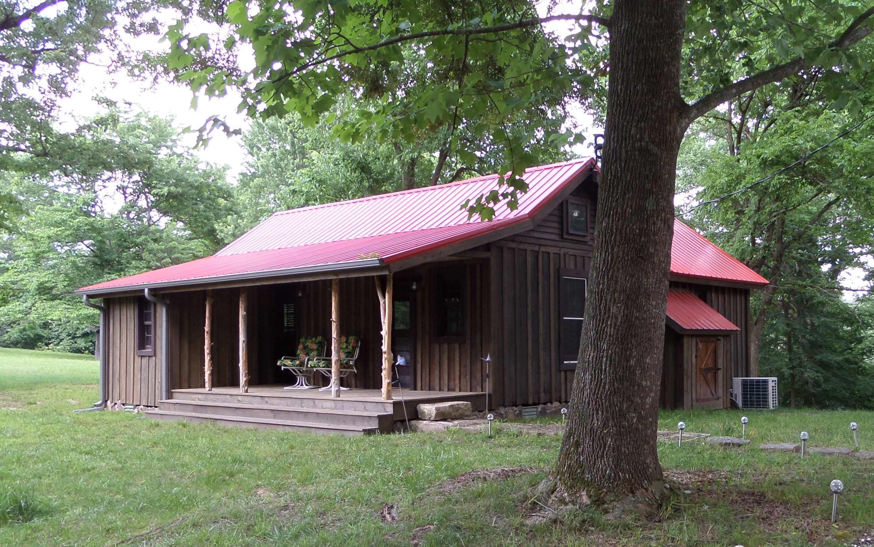 A rustic wooden cabin with a red roof, a porch with chairs, and a large tree in front.