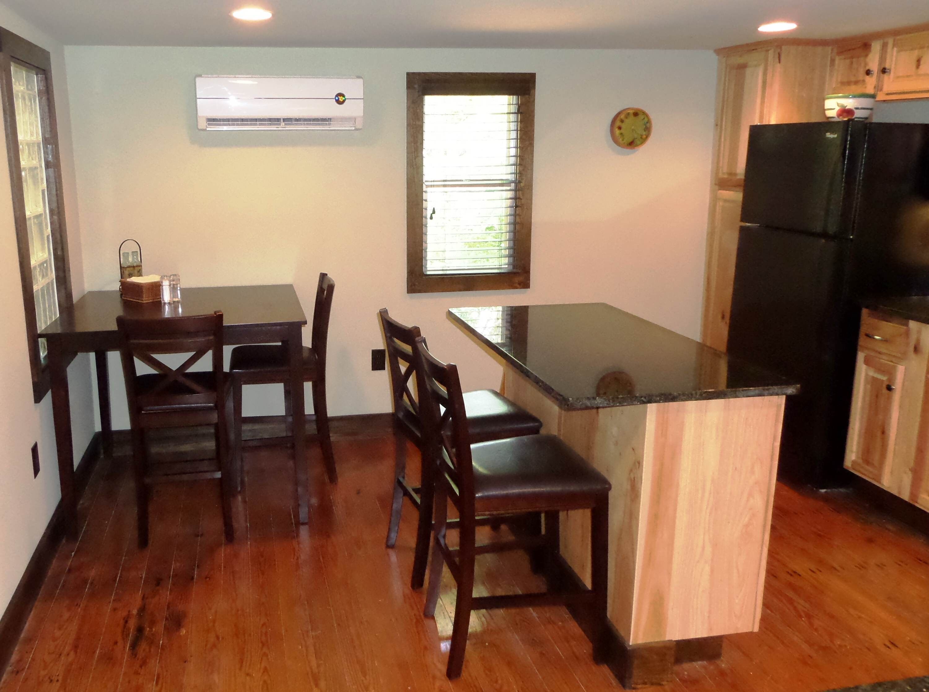 A kitchen with wooden cabinets, a black refrigerator, a dining table with chairs, and an air conditioning unit on the wall.