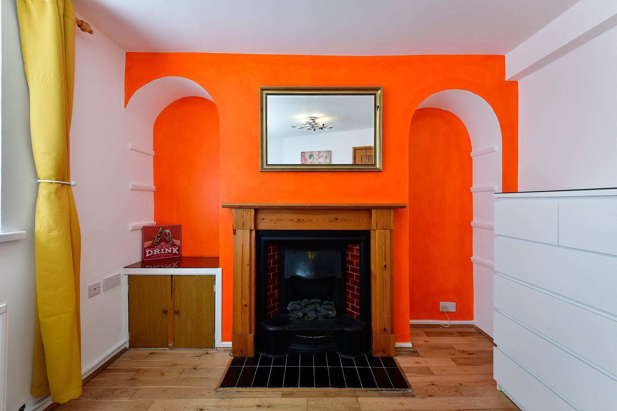 A room with an orange wall, a fireplace with a mirror above it, and a yellow curtain.