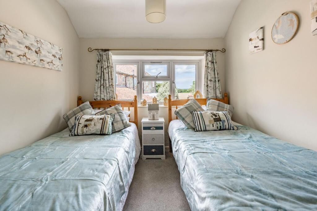 A bedroom with two beds, a window with curtains, a bedside table, and a wall-mounted clock.