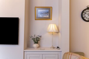 Una sala de estar con chimenea blanca, un cuadro colgado encima, un reloj en la pared y una lámpara en el repisa.