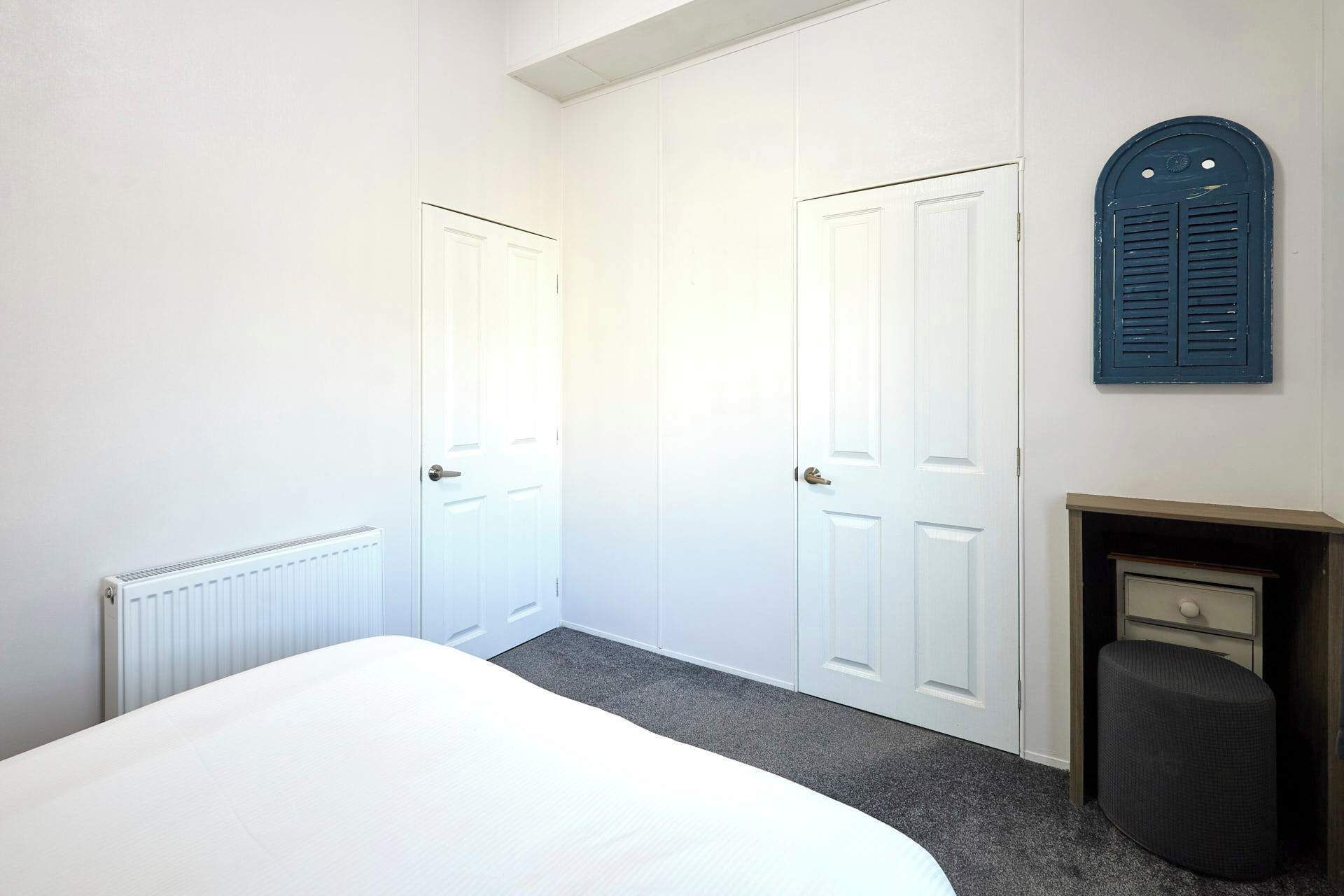 Een slaapkamer met twee witte deuren, een radiator, een klein openhaardvuur en een donkere poef.