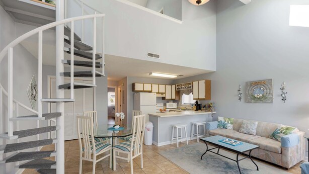 Ein modernes Interieur mit einer Wendeltreppe, einer Küche mit weißen Schränken, einem Essbereich mit Glastisch und einem Sofa mit gemustertem Überwurf.
