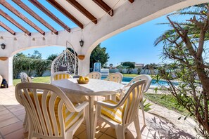 A covered patio with a pool, white chairs, and a table with yellow cushions.