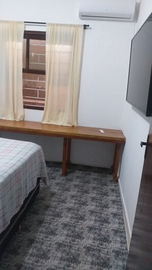 A room with a bed, a bench, a window with curtains, a wall-mounted air conditioner, and a door.