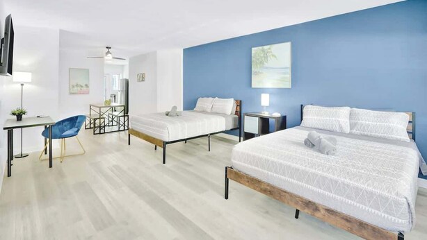 A hotel room with two beds, a desk, a chair, a TV, and a painting on the wall. - 1BR Miami Beach Apt w/ King + 2 Queens & Smart TV (Miami Beach)