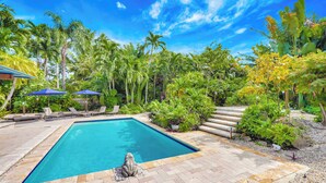 A rectangular swimming pool with a stone border, surrounded by lush greenery and palm trees. - Casa in Tropic Paradise with Pool and BBQ (Biscayne Park)