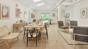A modern dining area with a long table, chairs, and a sofa. There are framed pictures on the walls and a window with a view of greenery. - Casa in Tropic Paradise with Pool and BBQ (Biscayne Park)