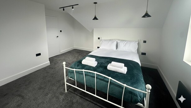 Een slaapkamer met een wit bed, groene beddengoed en een wit hoofdeinde. Twee zwarte plafondlampen en een raam aan de rechterkant.