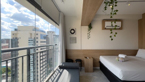 A modern hotel room with a large balcony, a bed, a bedside table, a small wooden nightstand, and a hanging plant. - Vitrali Moema (São Paulo)