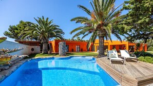Ein Schwimmbecken mit Holzterrasse und Liegestühlen, umgeben von Palmen und orangenen Gebäuden.
