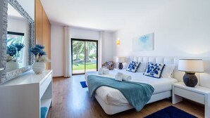 Ein modernes Schlafzimmer mit einem großen Bett, weißen Wänden und Blick auf einen Garten durch eine Schiebetür.
