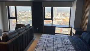 A modern living room with large windows, a sectional sofa, a bed with a striped bedspread, and a view of a cityscape. - Loft 1 Boudica, Puebla (San Bernardino Tlaxcalancingo)