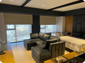 Une chambre d’hôtel moderne avec un canapé d’angle, un lit recouvert d’une courtepointe à carreaux, un bureau avec un ordinateur portable et de grandes fenêtres donnant sur la ville.