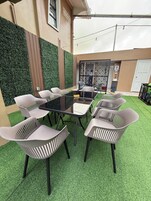 Un patio con una mesa de vidrio rodeada de sillas blancas de plástico, una cerca decorativa de metal y una pared cubierta con césped artificial verde.