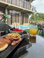 Una mesa de desayuno con huevos, tocino, tostadas, sandía y jugo de naranja. Se ve una piscina al fondo.